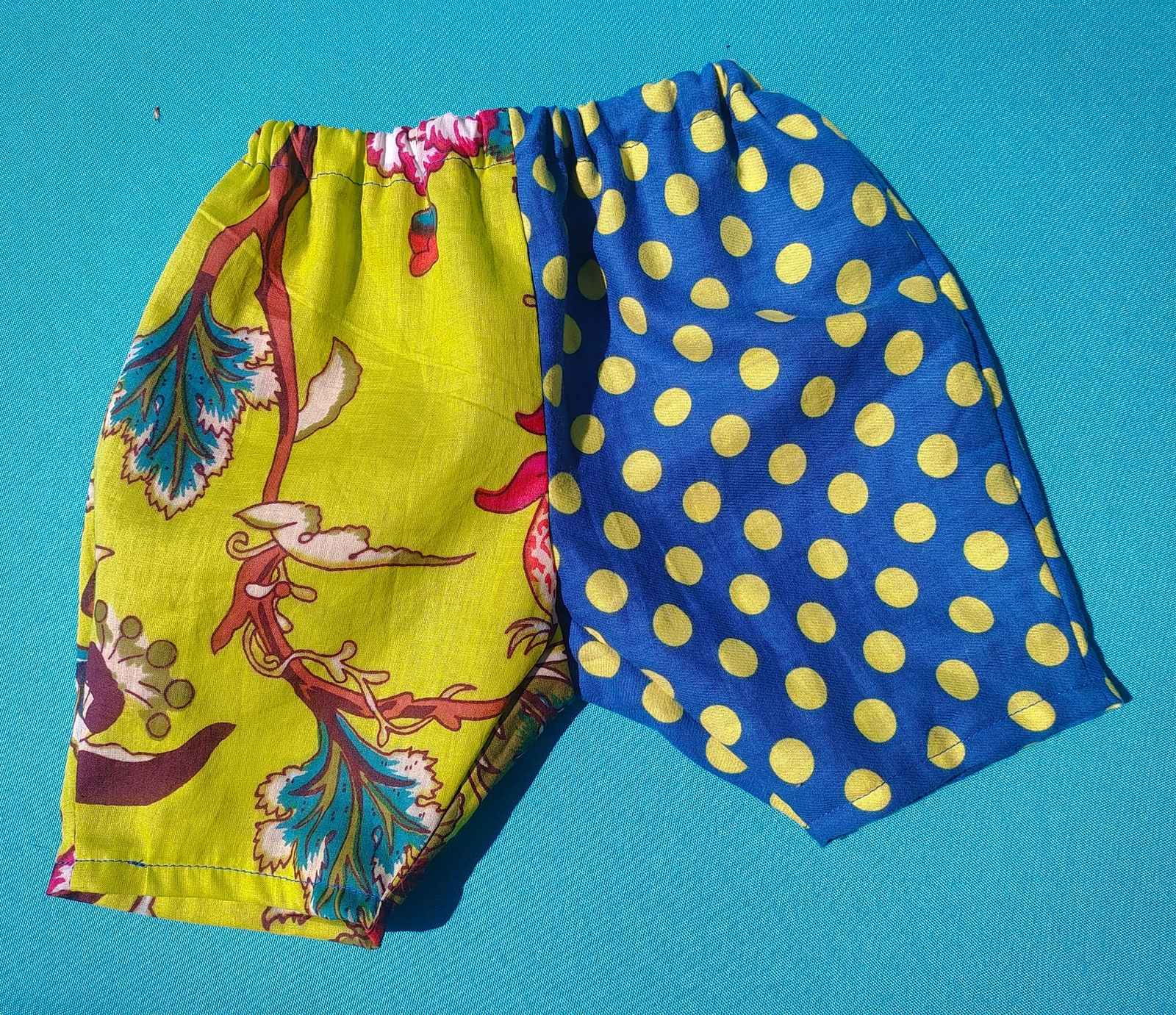 A tiny pair of baby pants laid flat on a turquoise background — one leg in chartreuse floral fabric, the other in blue with yellow polka dots